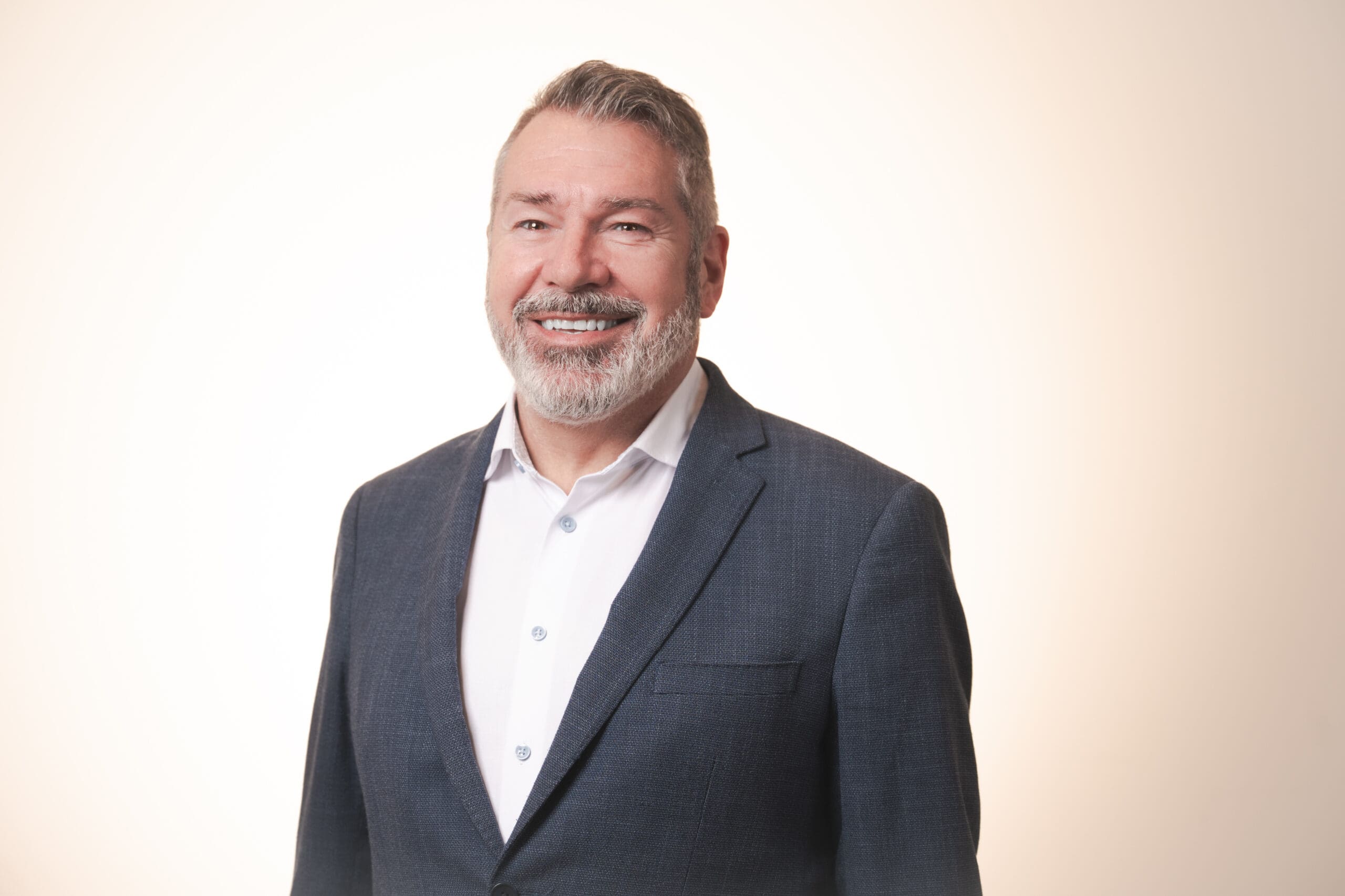 A middle-aged man with grey hair and a beard, wearing a dark suit jacket over a white shirt, smiles at the camera against a plain, light-coloured background.