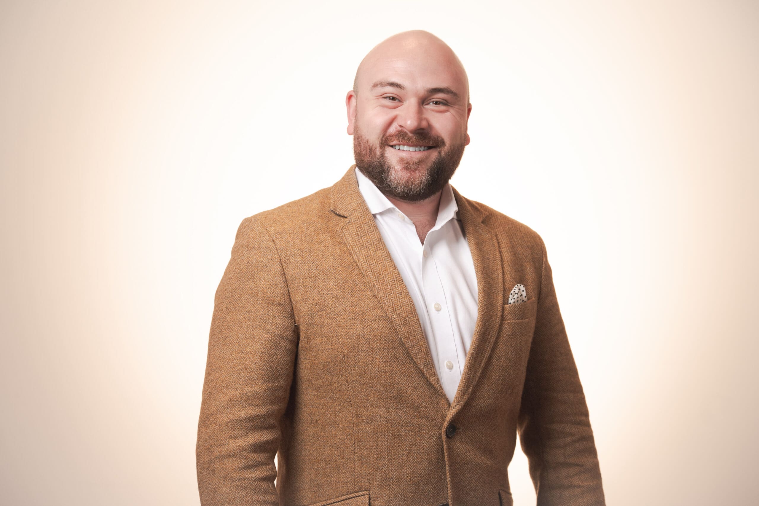 A smiling man with a bald head and beard wearing a brown blazer, white dress shirt, and patterned pocket square stands against a plain, light background.