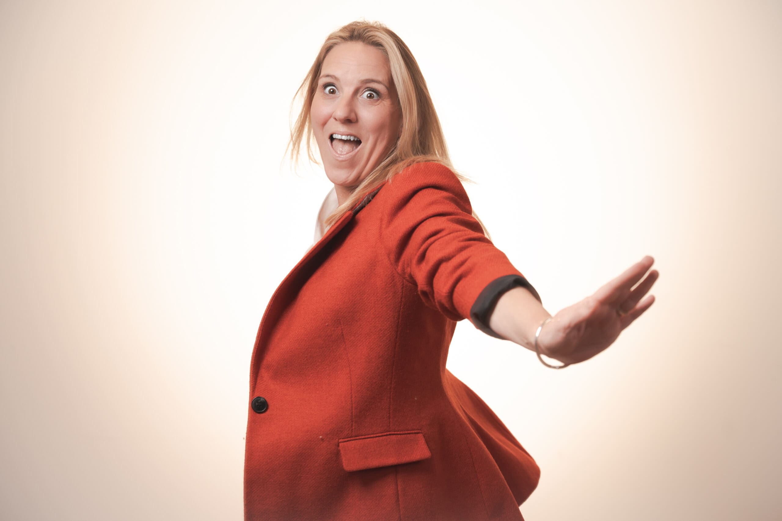 A woman wearing a red coat smiles widely whilst extending one arm backwards, as if in a playful or dancing motion, against a light-coloured background.