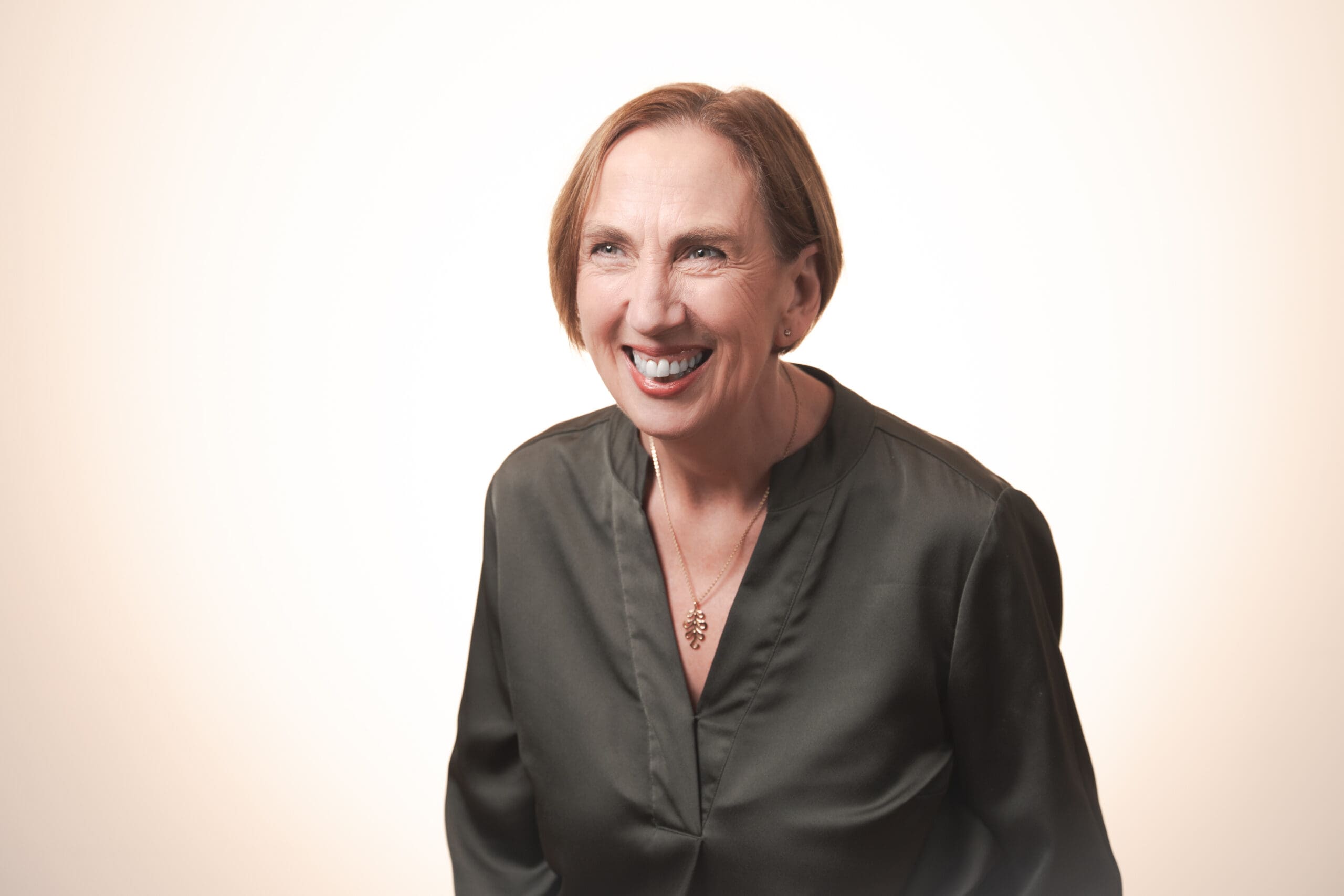 A middle-aged woman with short light brown hair, wearing a dark blouse and a pendant necklace, smiles broadly against a plain, light background.