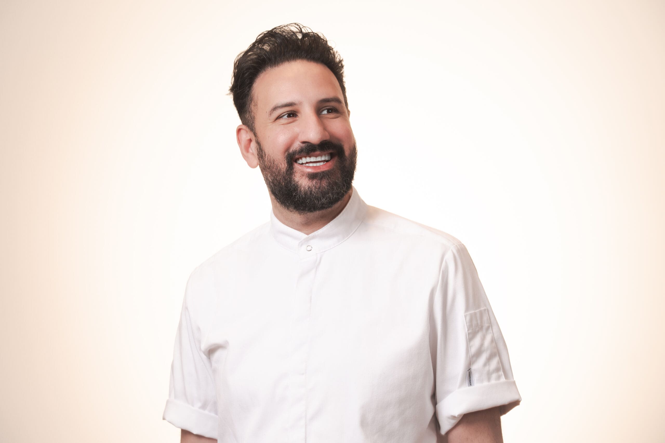 A smiling man with dark hair and a beard, wearing a white buttoned shirt, stands against a plain, light background.