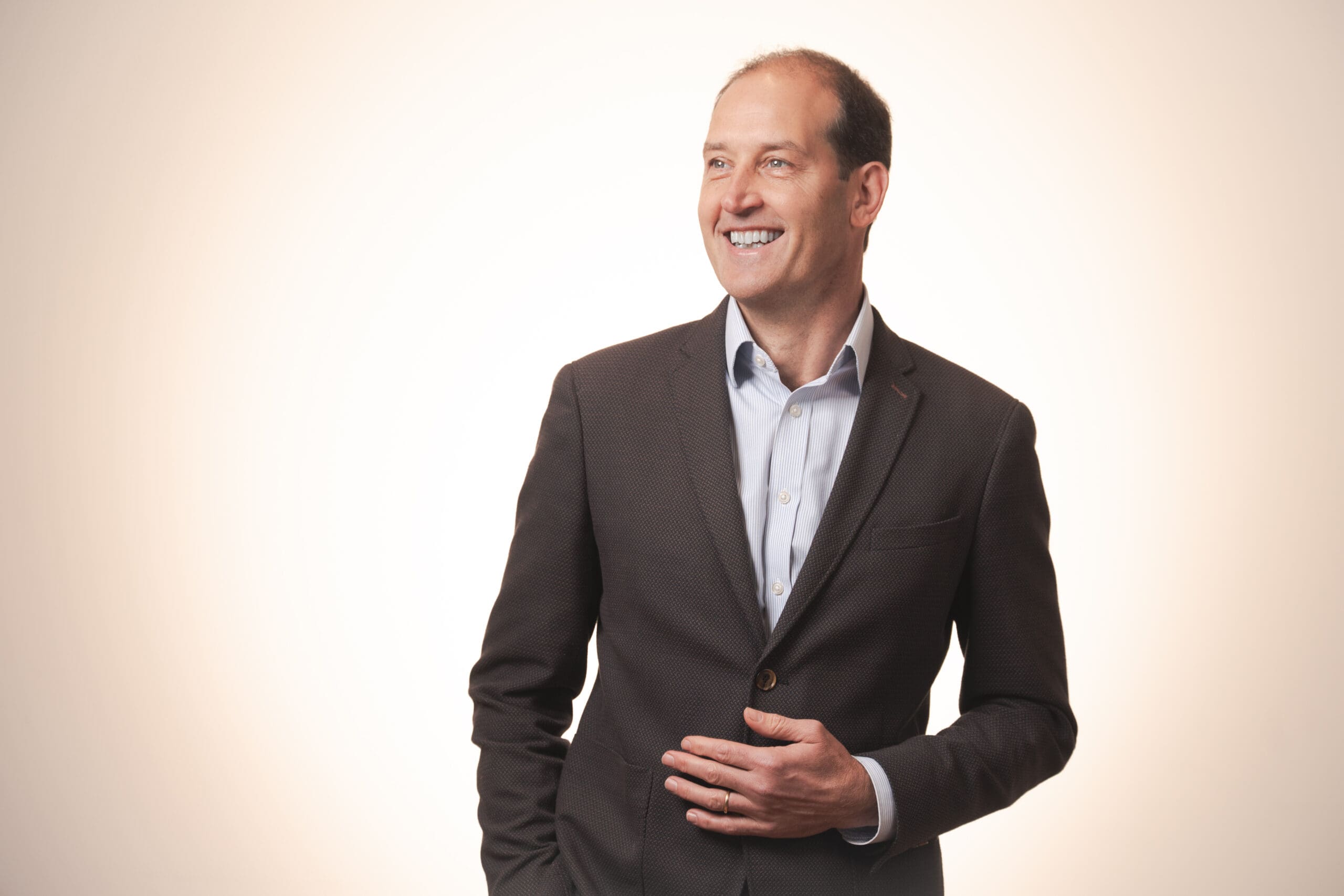 A man in a dark suit and light shirt stands smiling and looking to his left against a plain, light background. He has short hair and one hand resting near his waist.