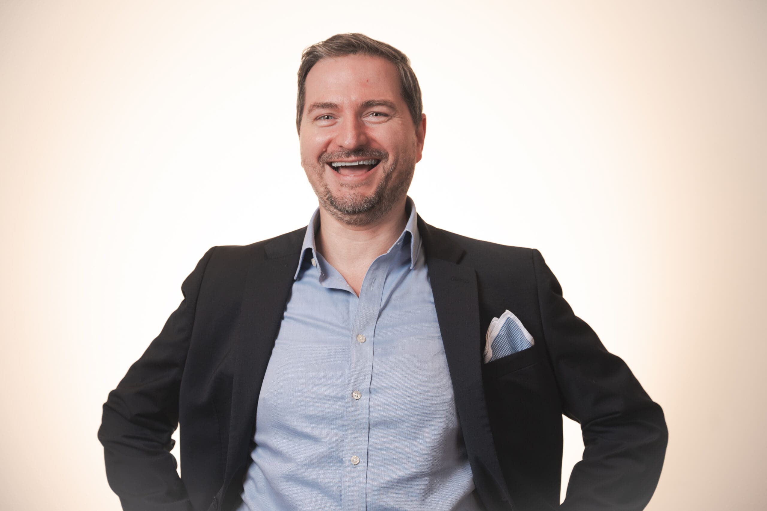 A man in a suit jacket and light blue shirt smiles broadly with his hands on his hips, standing against a plain, light background.