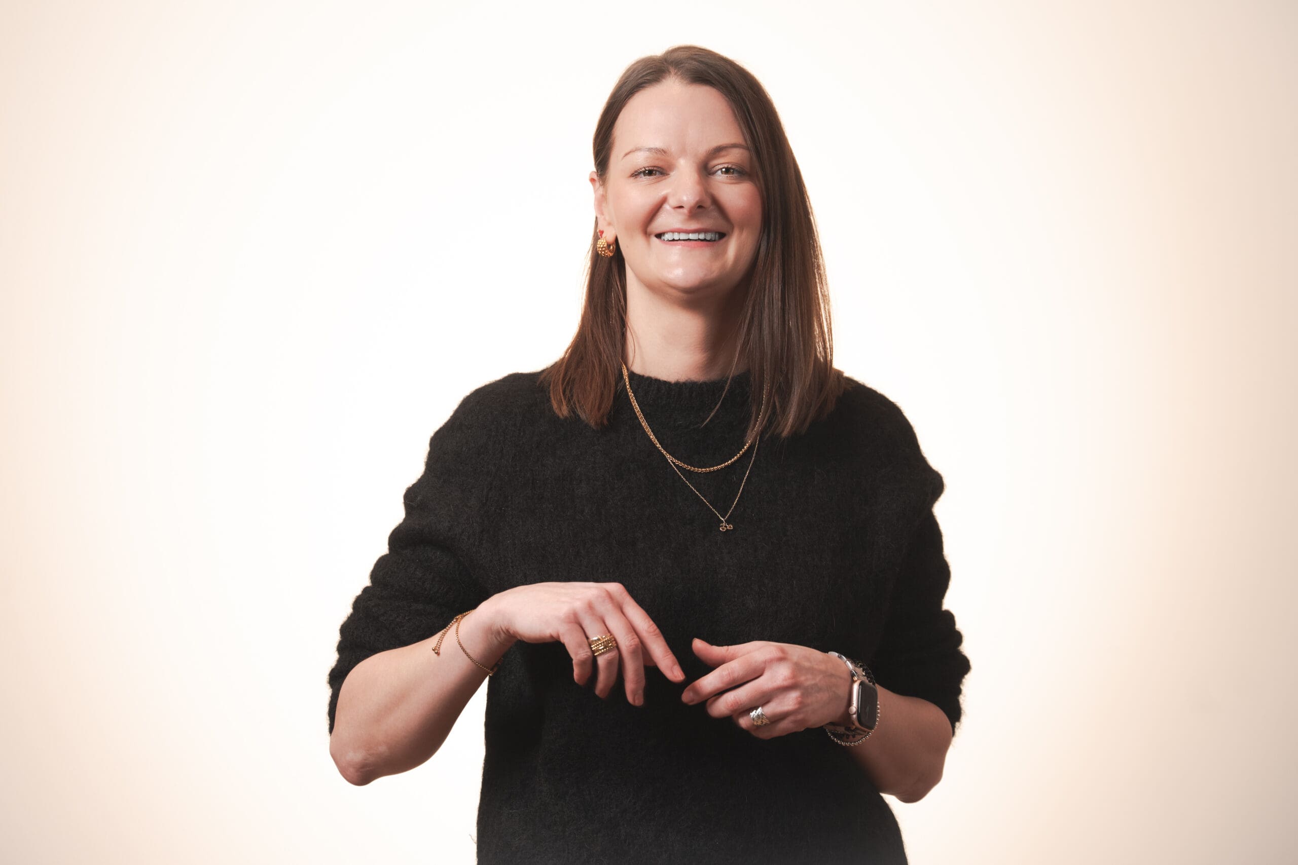 A woman with straight brown hair wearing a black jumper and layered necklaces smiles at the camera against a plain, light background. Her hands are visible in front of her, and she has a relaxed, friendly expression.