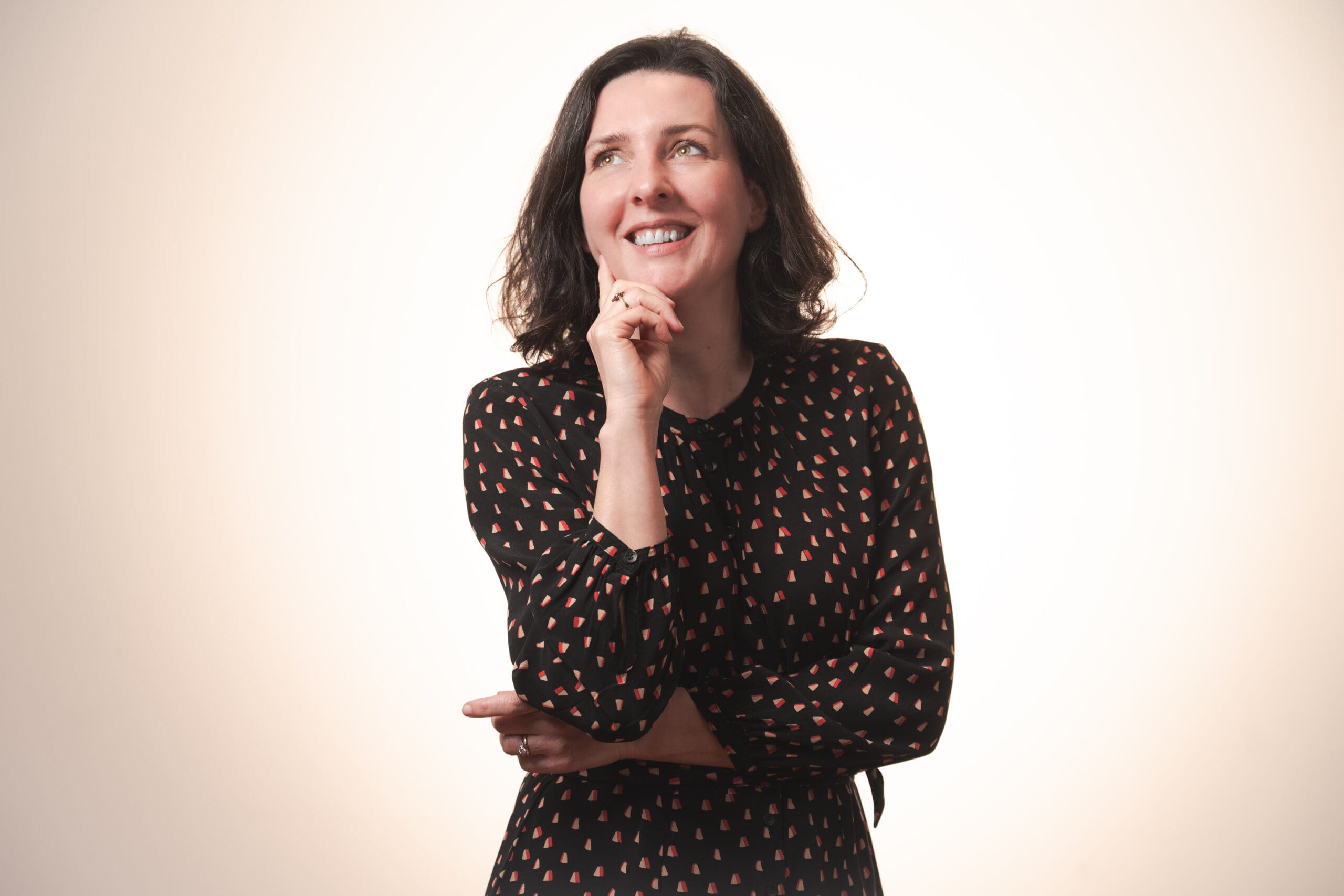 A woman with shoulder-length dark hair wearing a black dress with a colourful pattern stands smiling and looking upwards, resting her chin on her hand in a thoughtful pose against a light background.