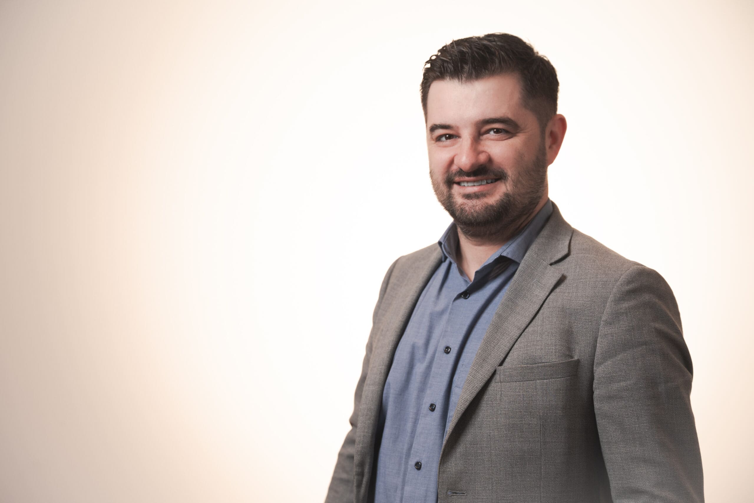 A man with short dark hair and a beard is smiling while wearing a grey blazer over a blue button-up shirt, standing against a plain, light-coloured background.