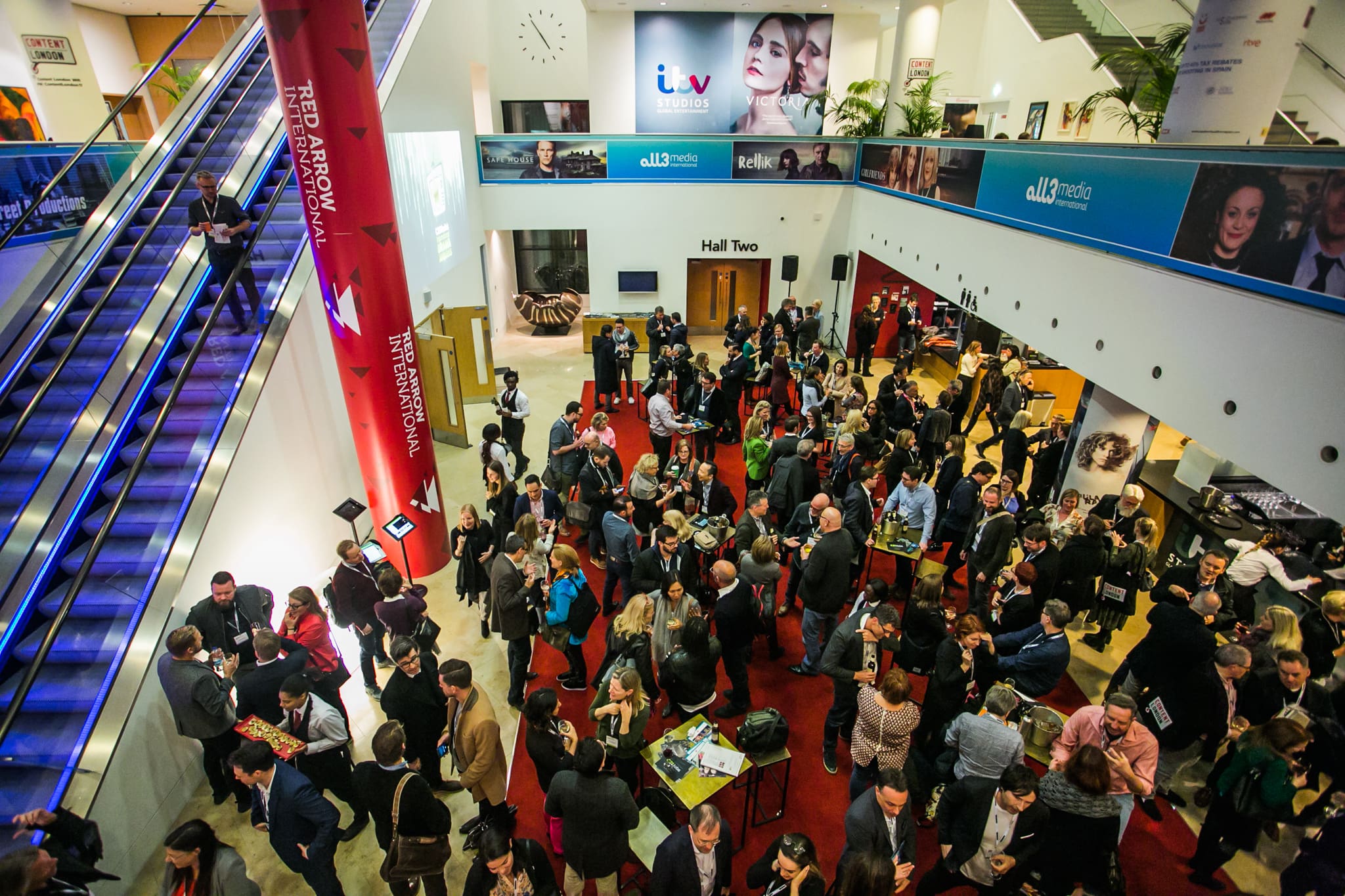 A large crowd of people mingle and network in a brightly lit, modern lobby with red carpet, escalators, and TV company banners and adverts on the walls.