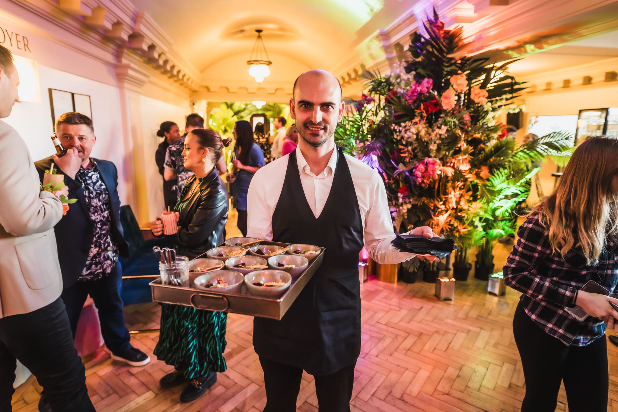 A waiter in a white shirt and black waistcoat holds a tray of appetisers at a lively indoor event with people socialising and a large, colourful floral arrangement in the background.