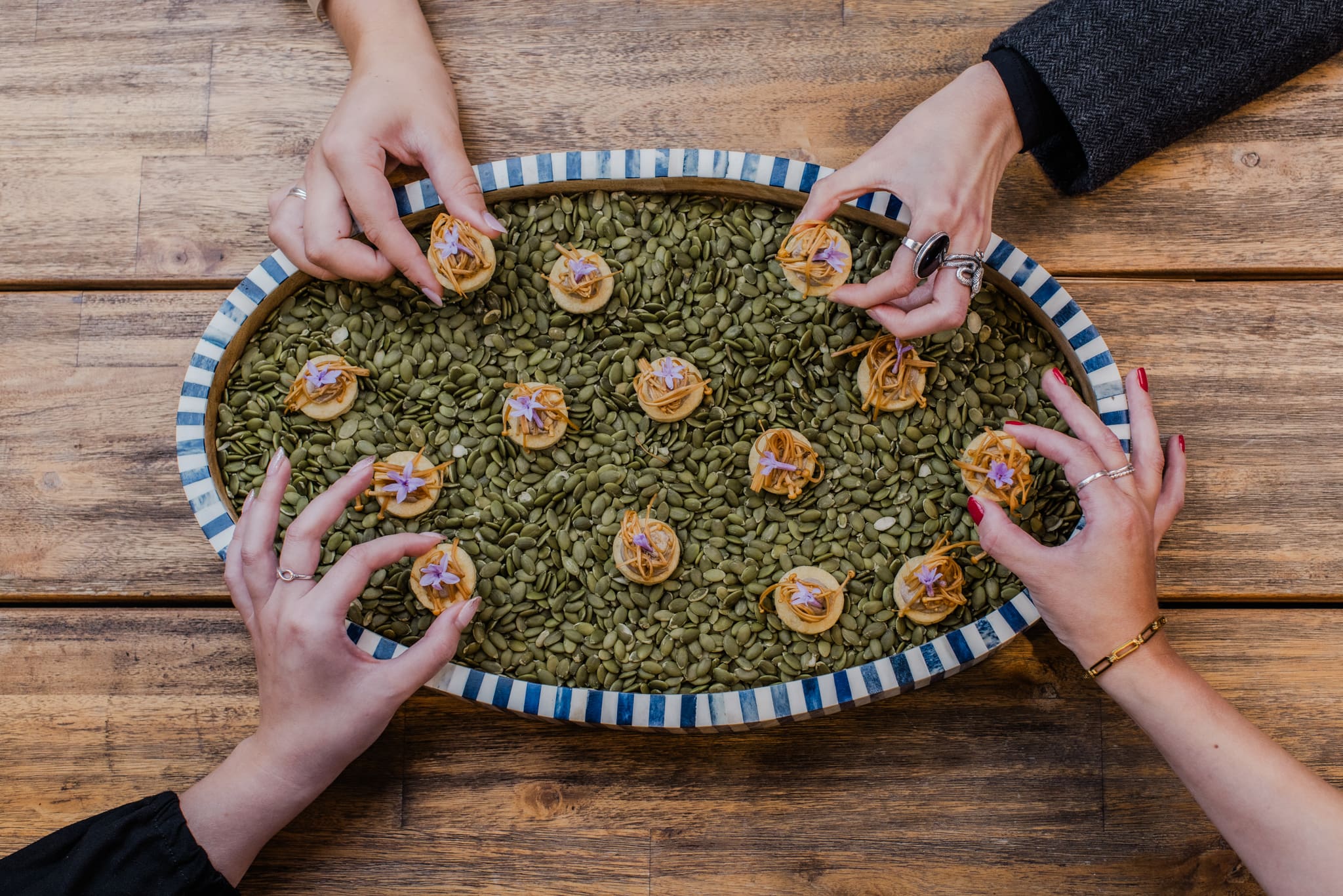 Three hands reach for small, artfully arranged appetisers topped with purple flowers, displayed on a striped platter filled with green pumpkin seeds, set on a wooden table.