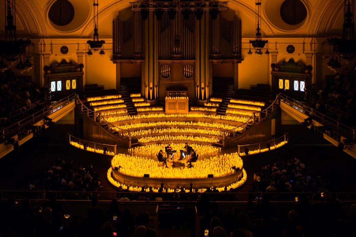A candlelit concert in a grand auditorium features musicians playing on a stage surrounded by glowing candles, with an audience seated in dimly lit rows. Warm yellow lighting creates a cosy, magical atmosphere.