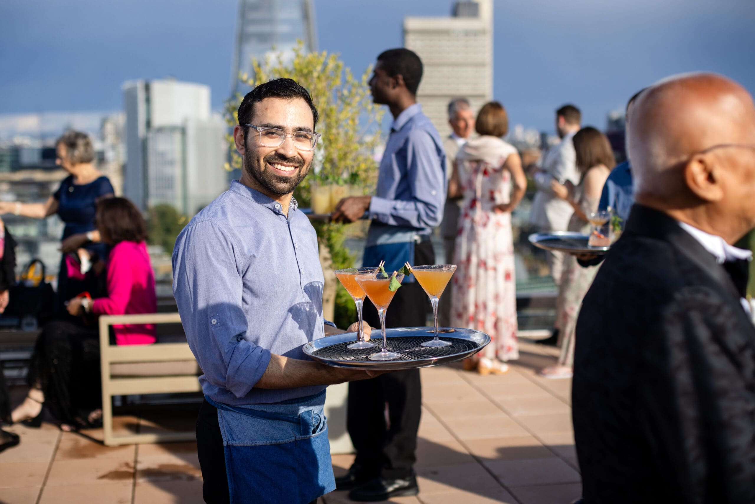 A smiling waiter holds a tray with colourful cocktails at an outdoor rooftop event with people socialising in the background and city buildings visible.