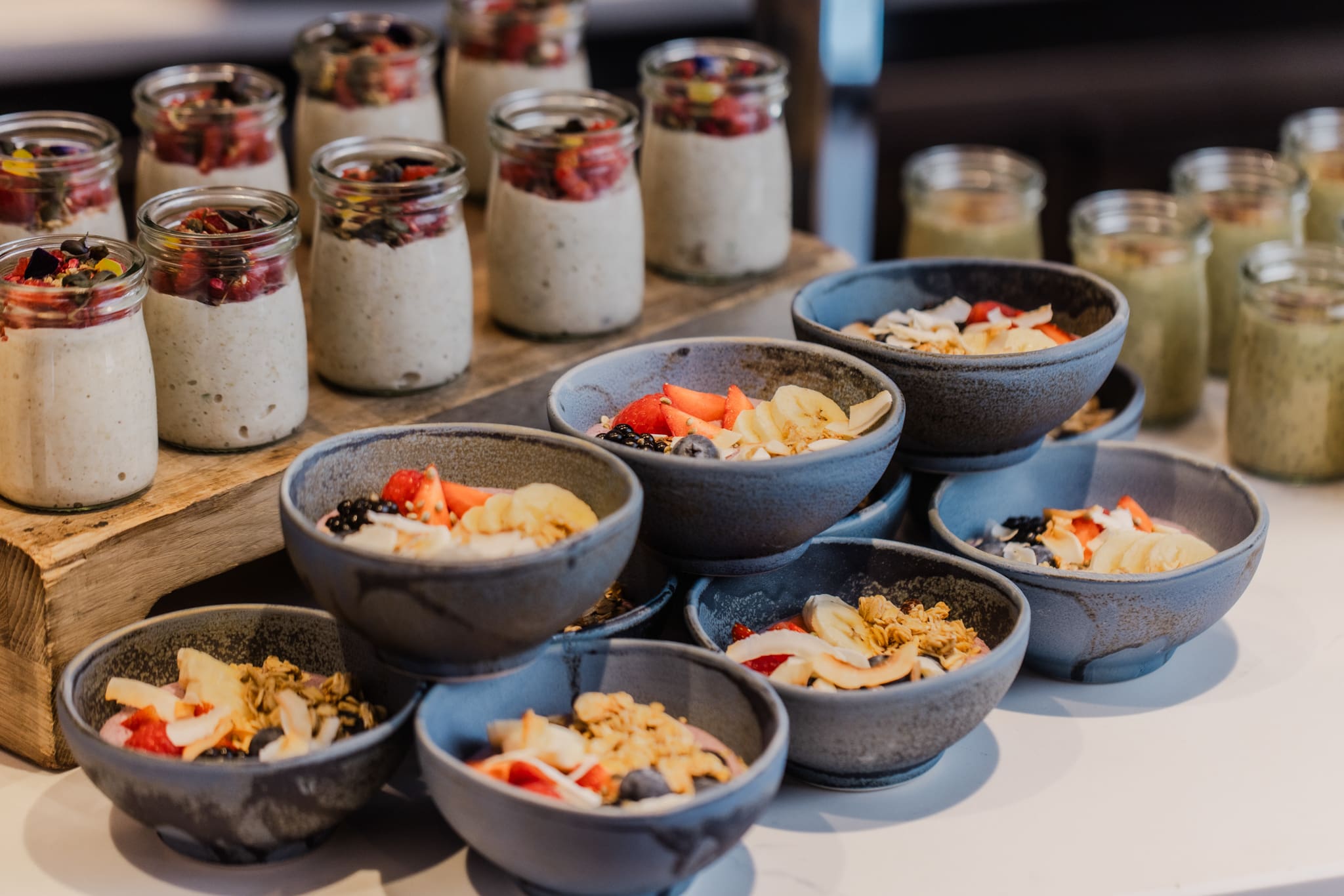 Small blue bowls filled with yoghurt, granola, and fresh fruit are stacked on a table. Behind them, jars of chia pudding topped with berries are arranged on a wooden tray.