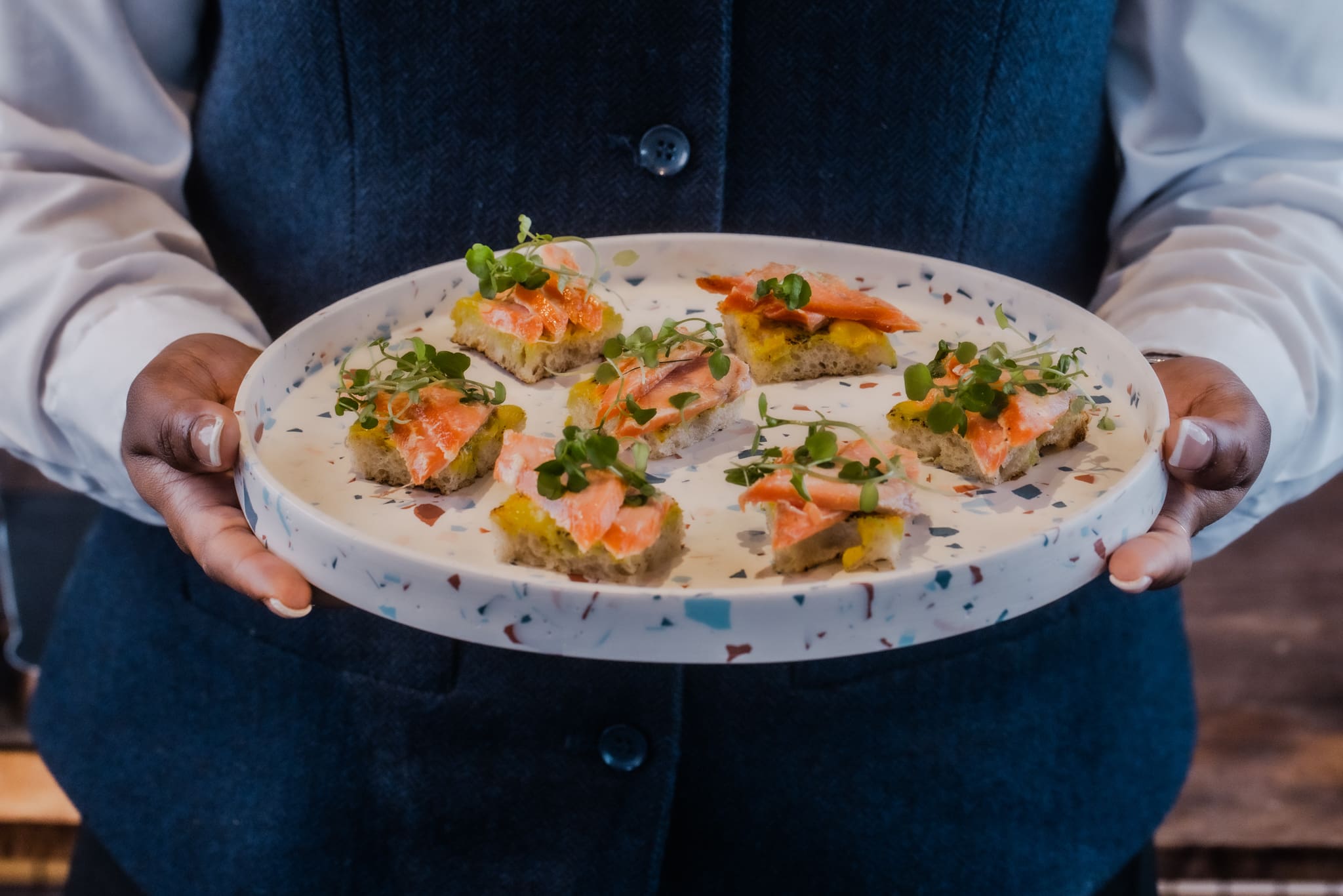 A person wearing a dark waistcoat and white shirt holds a round plate with six pieces of starter toast topped with salmon and microgreens.