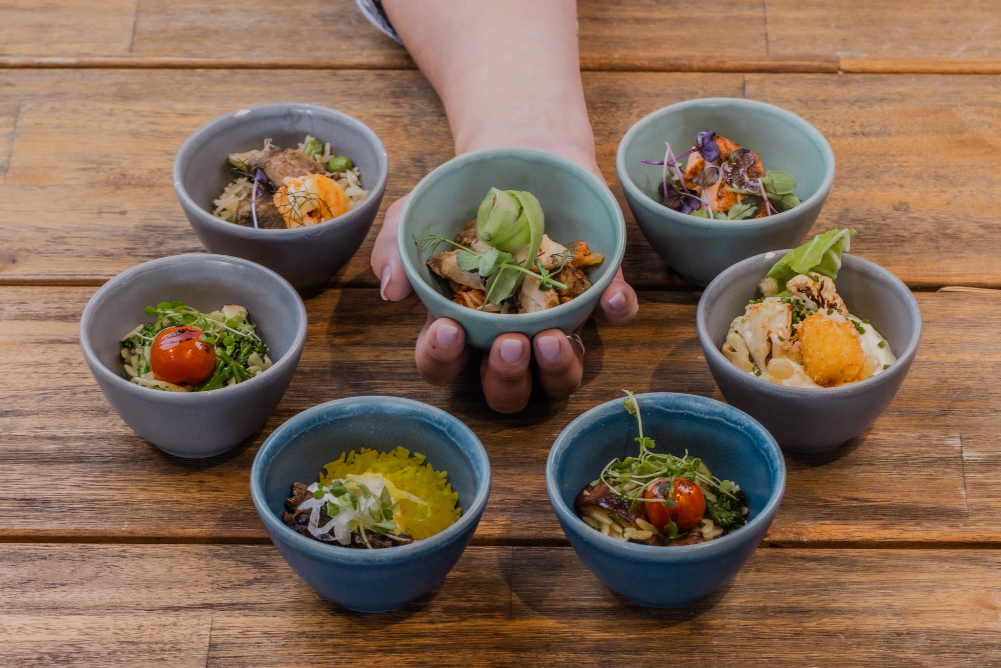 A hand holds a blue bowl with vegetables, surrounded by six other colourful bowls filled with assorted salads and garnished dishes, all arranged on a wooden table.