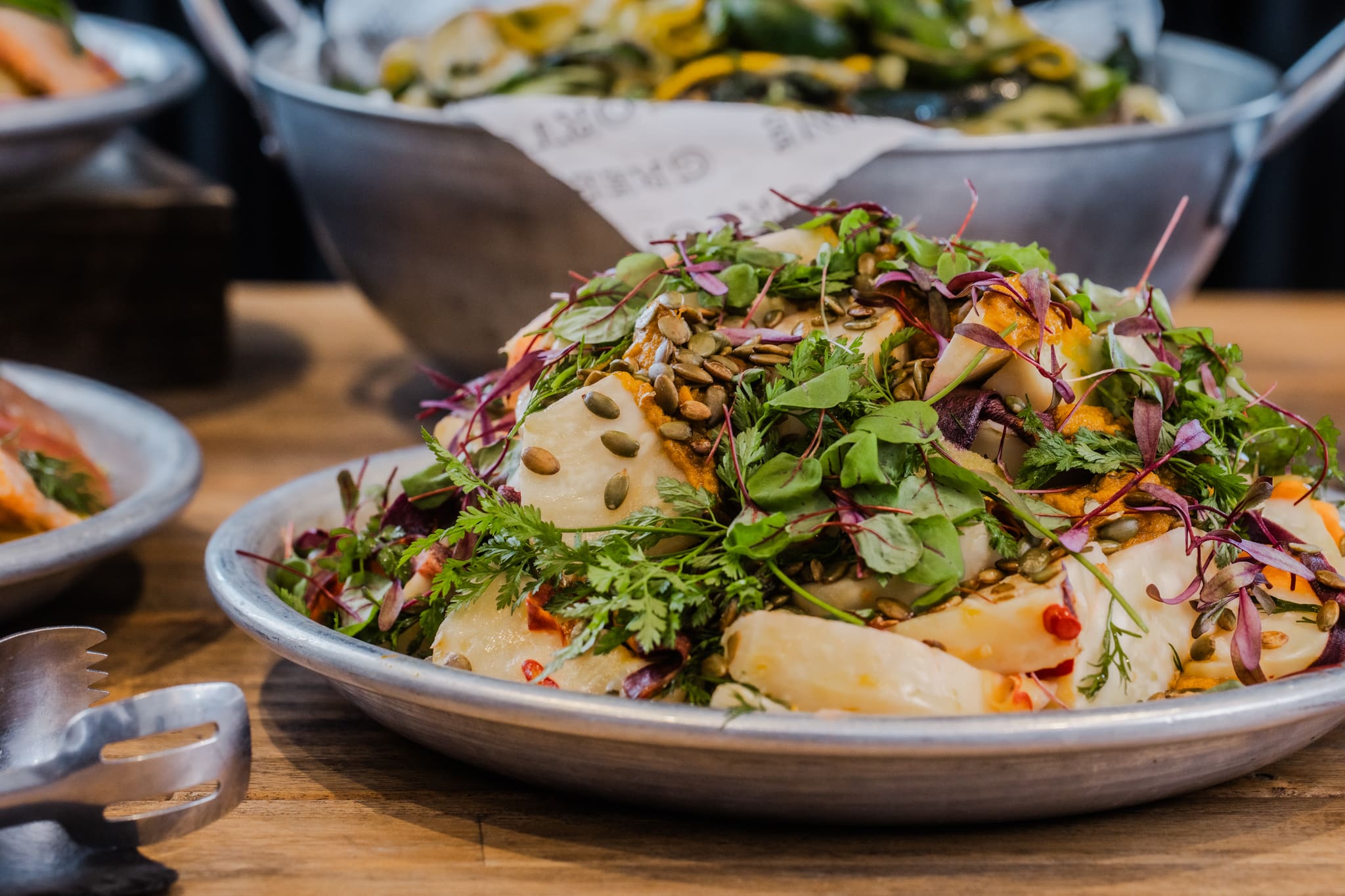 A metal plate filled with a colourful salad featuring sliced vegetables, leafy greens, herbs, and seeds, set on a wooden table with serving tongs beside it. Other dishes are visible in the blurred background.