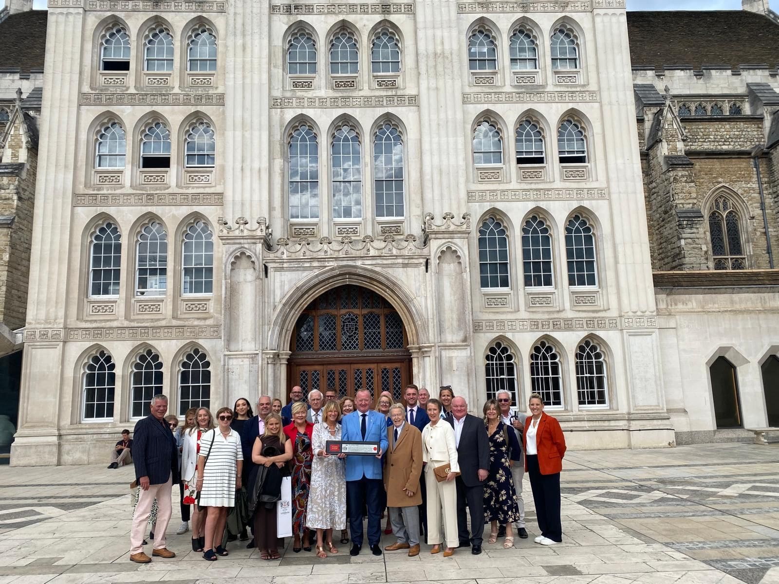 A group of people pose for a photo in front of a large historic building with arched windows and ornate stonework. Most are dressed formally and stand in a semicircle, with one person holding a rectangular plaque.