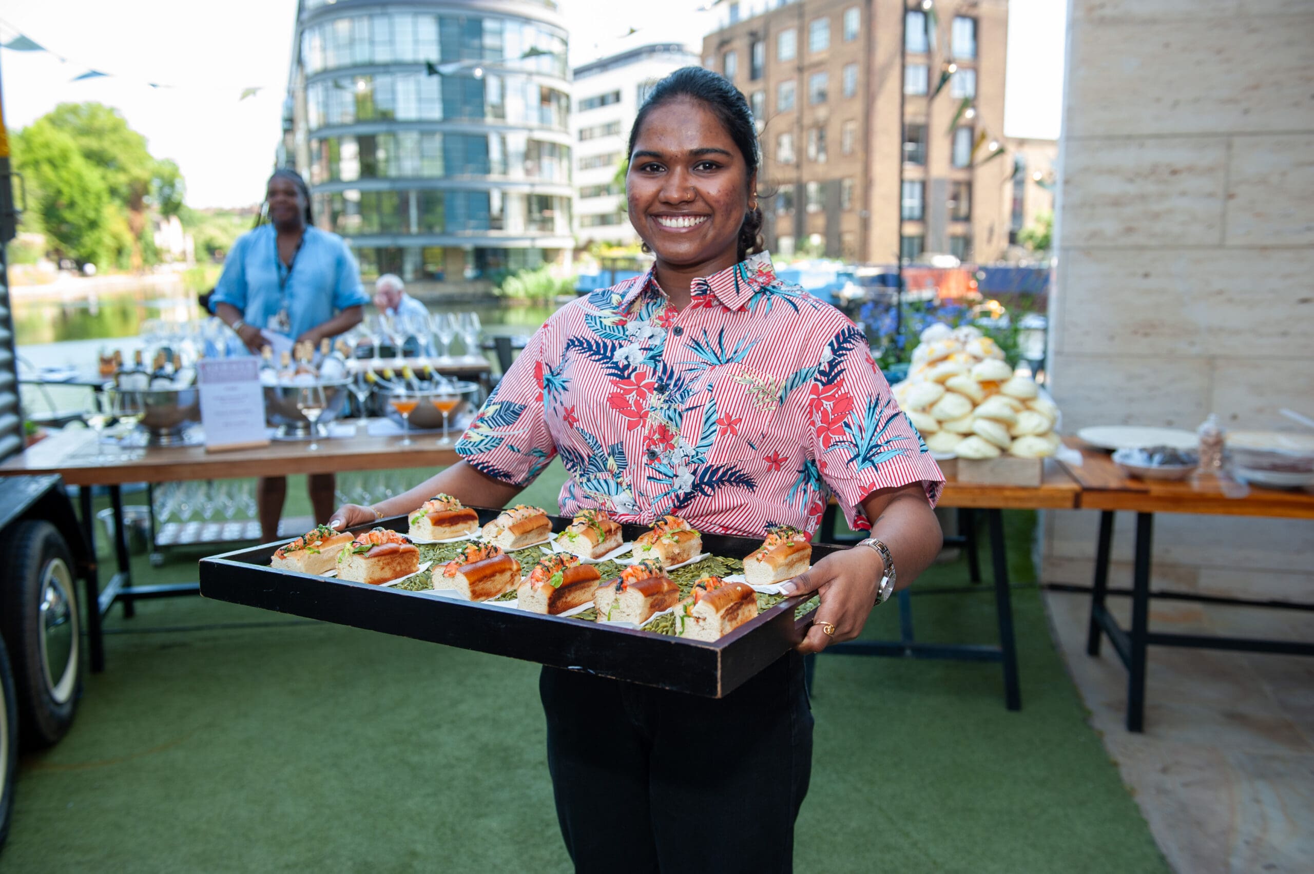 A smiling person in a floral shirt holds a tray of small sandwiches at an outdoor event, with tables of food and modern buildings in the background.