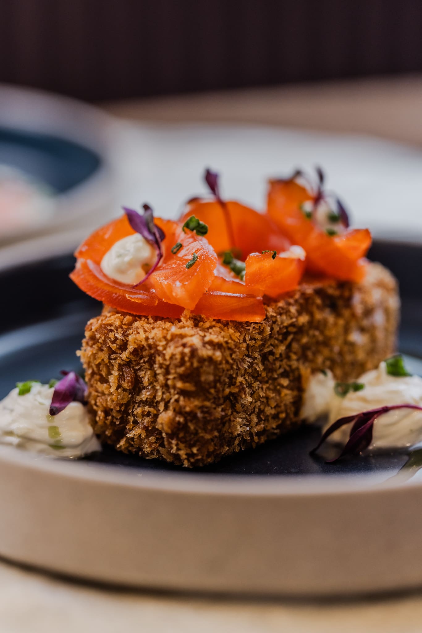 A close-up of a breaded and fried rectangular food item topped with slices of smoked salmon, dollops of cream, microgreens, and served on a black plate with additional cream and garnish on the side.