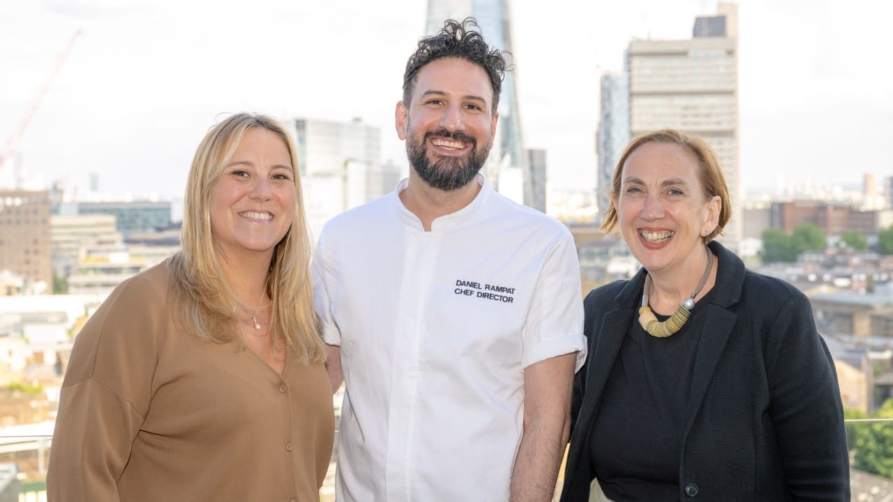 Three people stand smiling outdoors with a city skyline in the background. The man in the middle wears a white chef's jacket, while the two women on either side wear business casual attire.