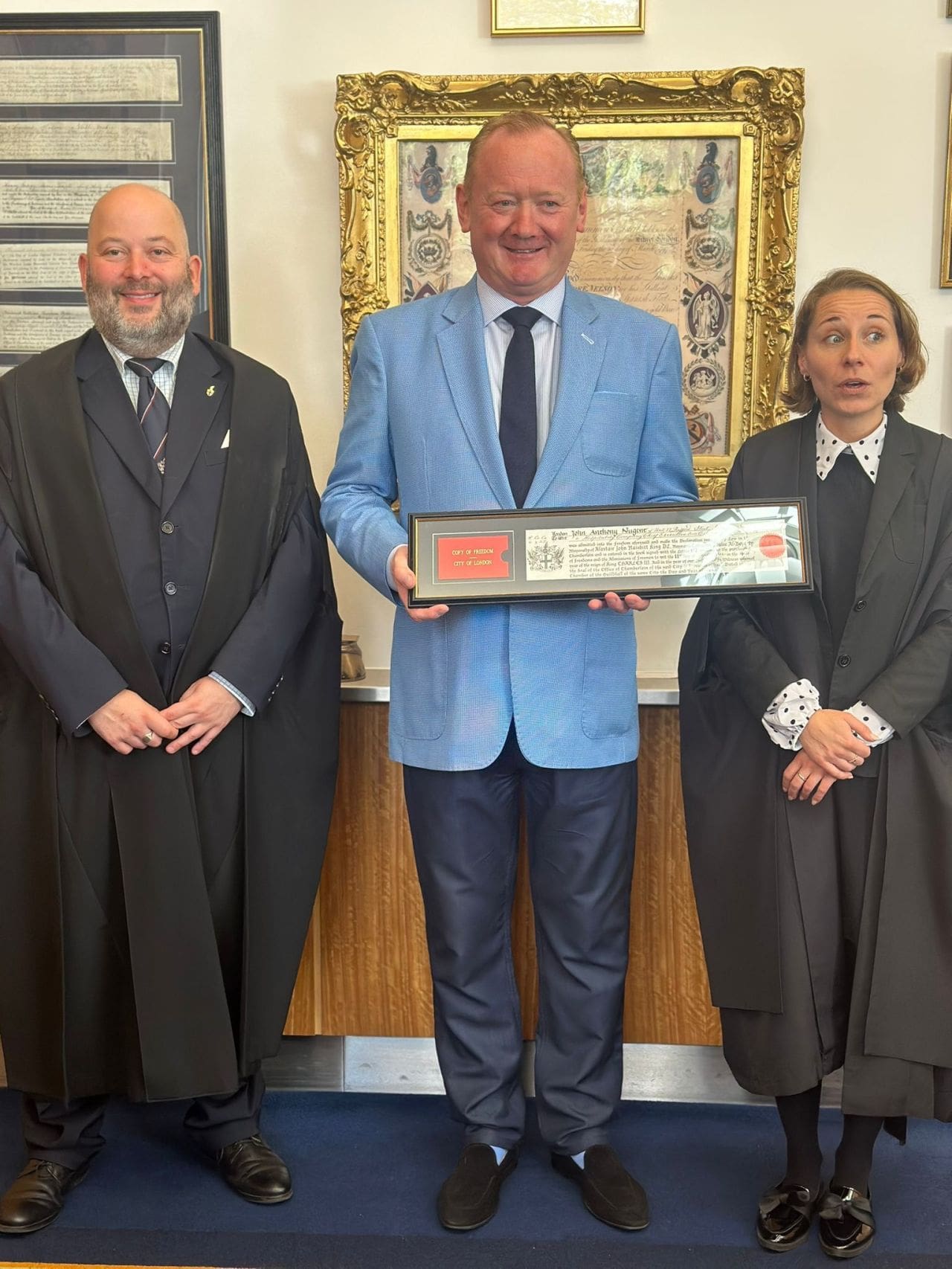 Three people stand indoors in formal attire. The central person, in a light blue jacket, holds a framed certificate and smiles. The two others, wearing black gowns, stand on either side, with a gold-framed document behind them.
