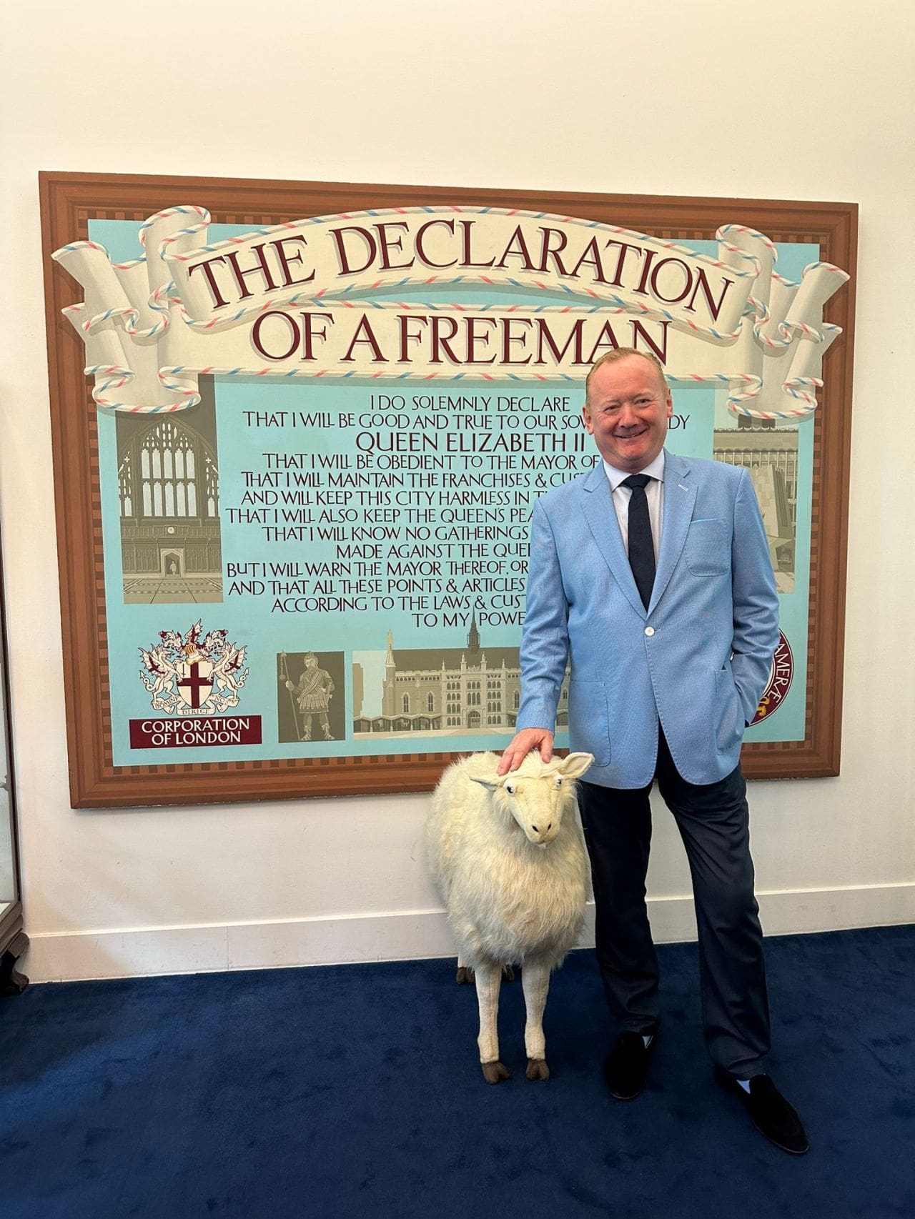 A man in a blue suit smiles whilst standing indoors next to a sheep. Behind them is a large sign reading “The Declaration of a Freeman” with other text and a crest.