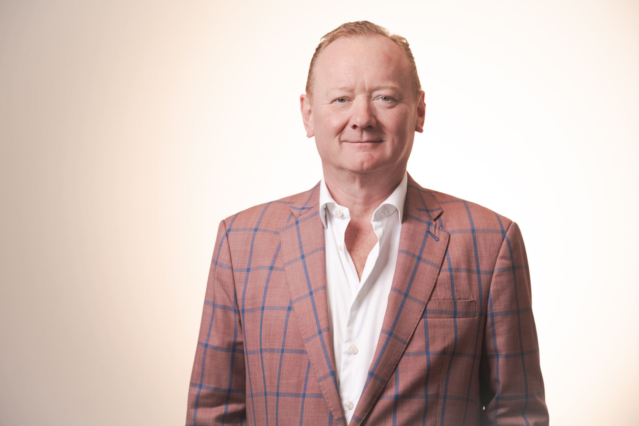 A middle-aged man with short fair hair wears a pink checked jacket over a white shirt, standing against a plain, softly lit background, smiling slightly at the camera.
