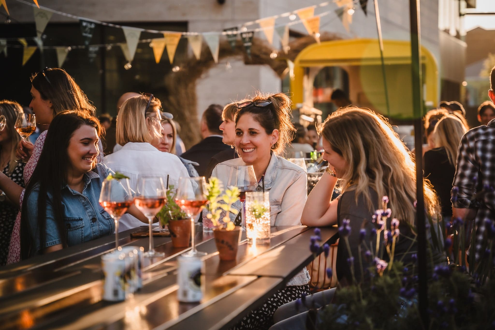A group of people sit at outdoor tables decorated with drinks and plants, chatting and smiling in warm sunlight. Festive bunting hangs above, creating a lively, cheerful atmosphere.