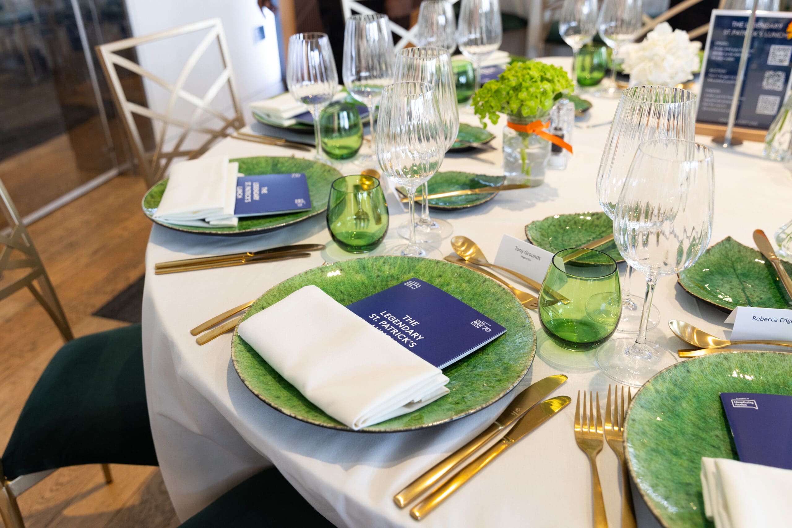 A formal dining table set with green plates, gold cutlery, folded white serviettes, green glasses, wine glasses, and navy blue booklets titled 