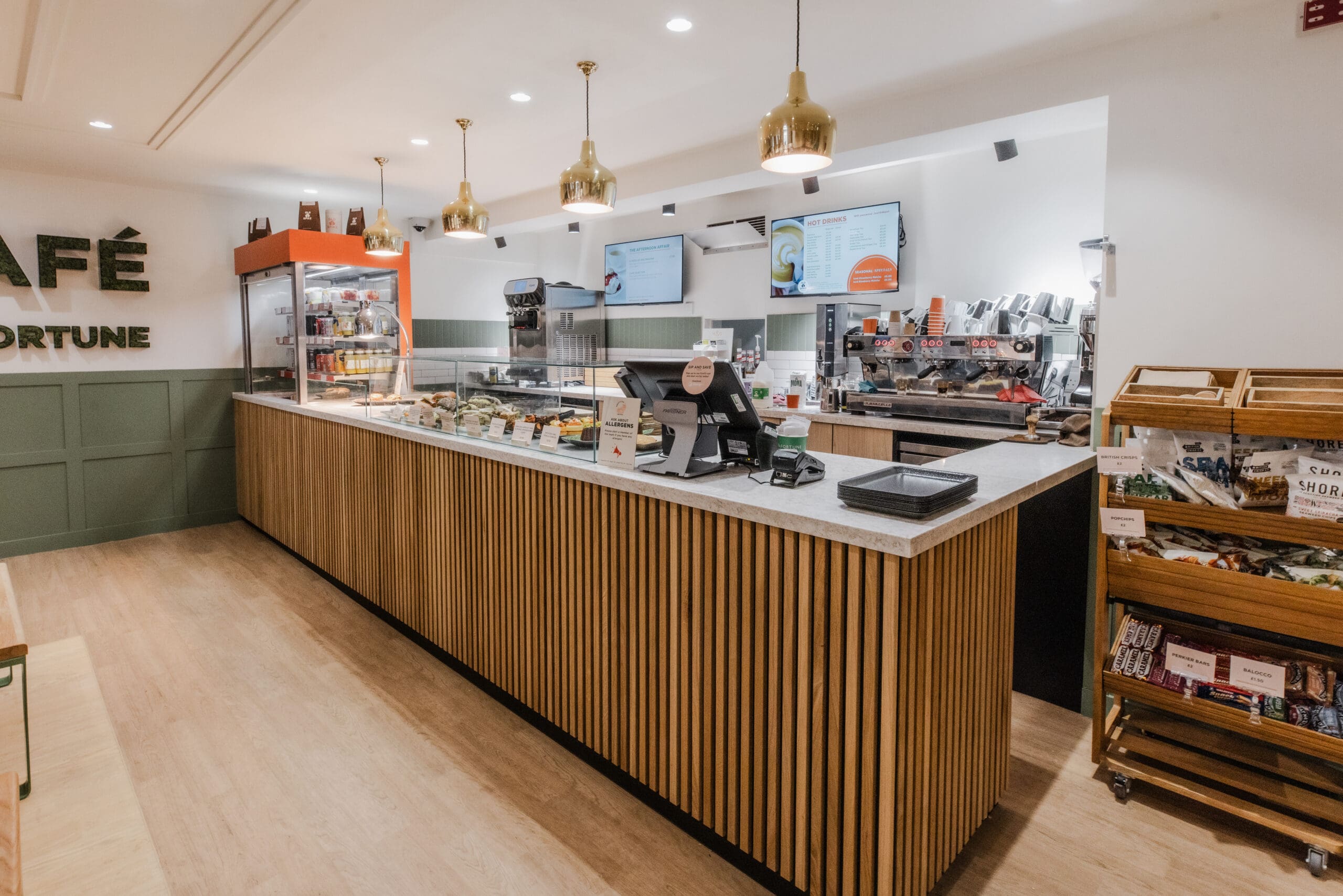 A modern café counter with a display case of pastries, two digital menu screens, coffee machines, and a snack rack on the right. The space has wooden accents and warm lighting.