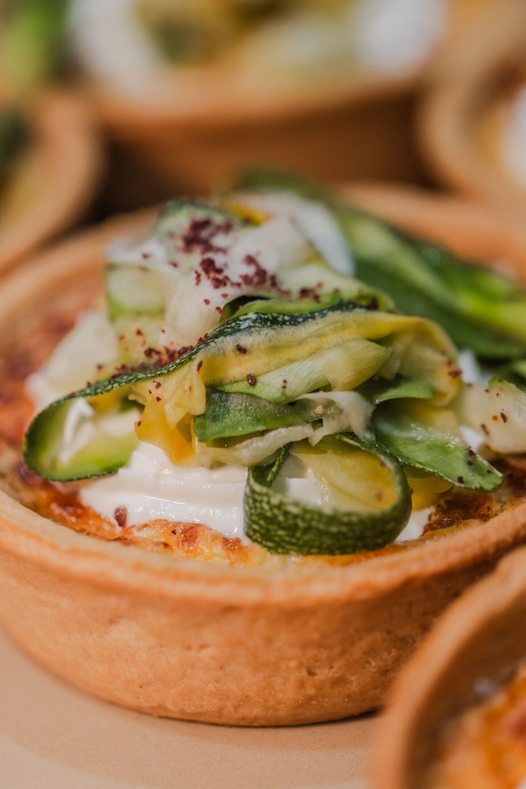 A close-up of a savoury tart with a golden crust, topped with creamy white cheese and thinly sliced, curled courgette, garnished with herbs.