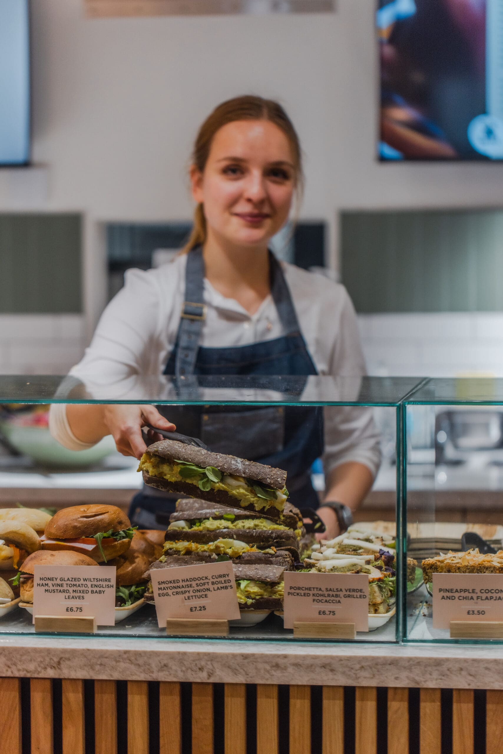 A woman in an apron stands behind a glass counter displaying assorted sandwiches, reaching to serve a stacked charcoal bread sandwich. Price and description cards are visible in front of each food item.