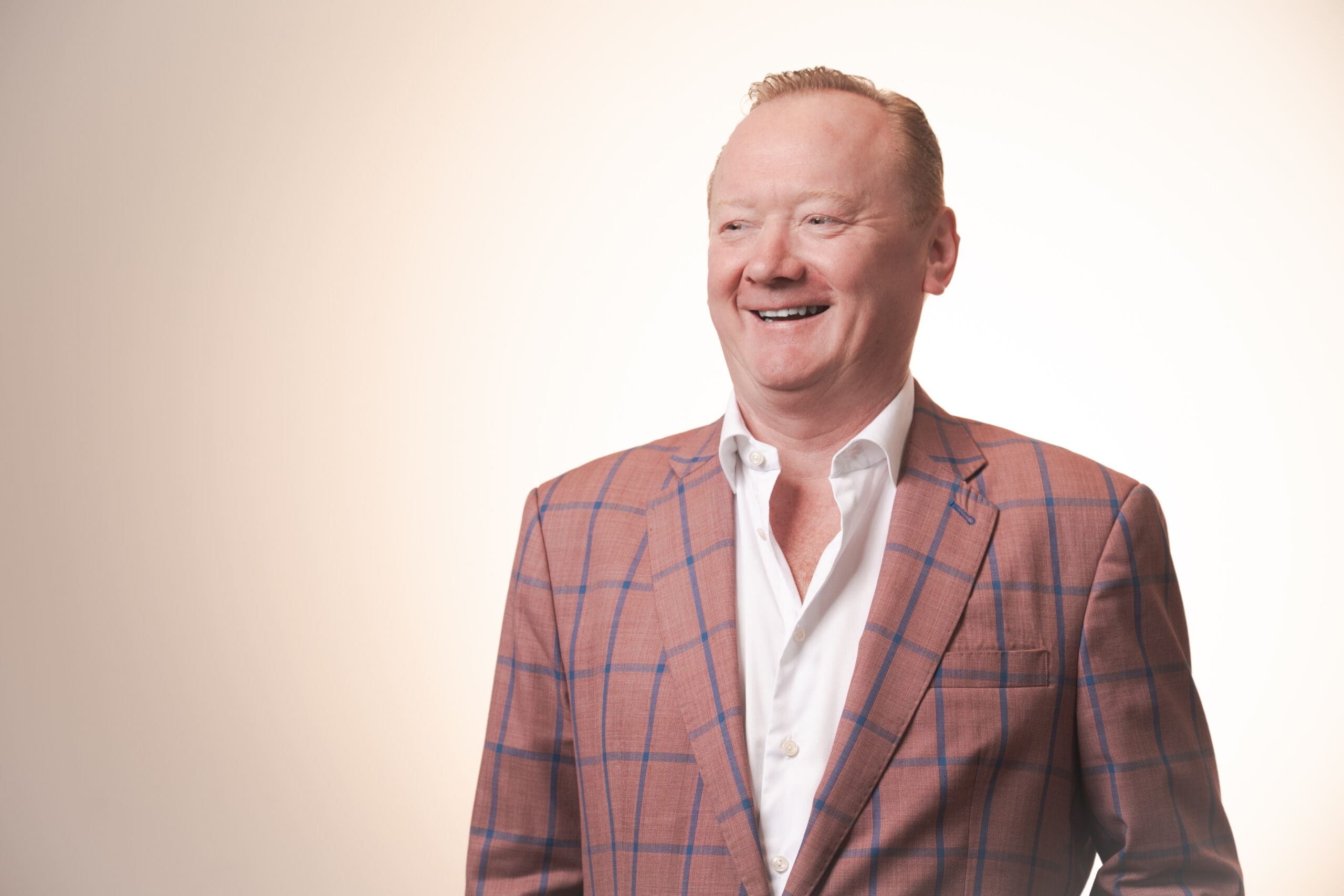 A smiling man with short fair hair wears a white dress shirt and a pink checked blazer. He stands in front of a light-coloured background, looking slightly to the side.