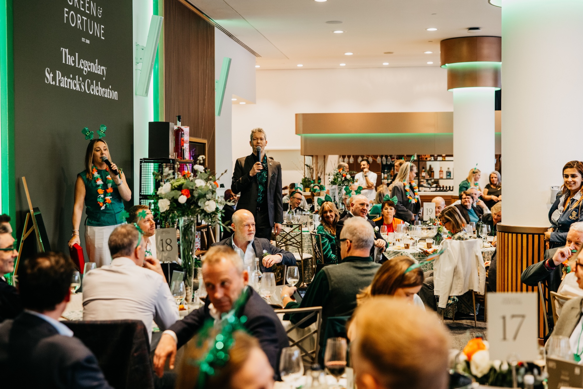 A lively indoor event with people seated at tables, some wearing green decorations for St. Patrick's Day. Two speakers stand at the front, one with a microphone, addressing the audience.