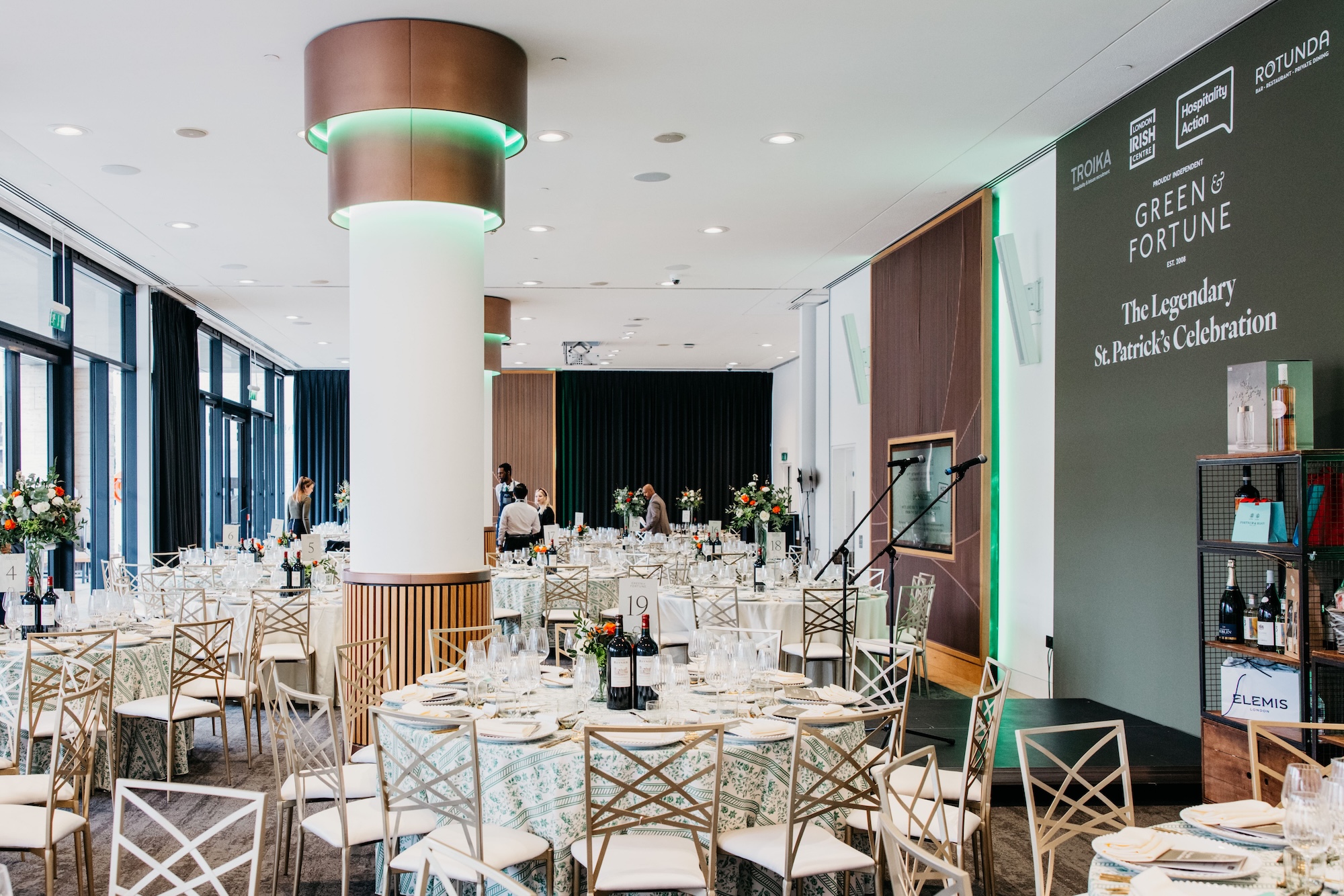 Elegant banquet hall set for an event, with round tables covered in patterned cloths, neatly arranged plates and glasses, floral centrepieces, and a large sign on the wall reading “Green & Fortune: The Legendary St. Patrick’s Celebration”.