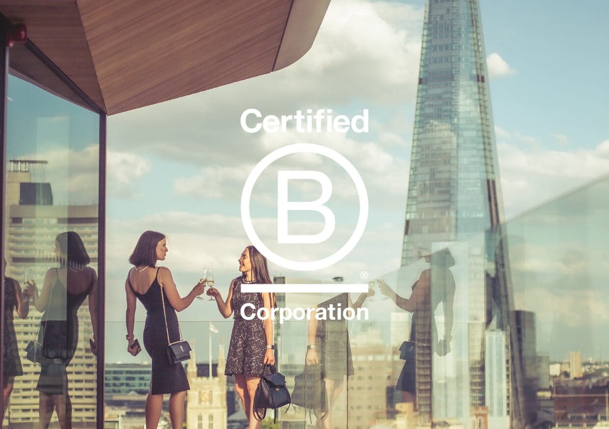 Two women in dresses stand on a rooftop terrace, toasting drinks, with The Shard and city buildings in the background. The 