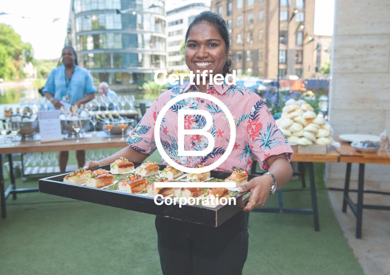 A smiling person holding a tray of food at an outdoor event, with tables of refreshments in the background. The B Corporation certification logo is overlaid on the image.