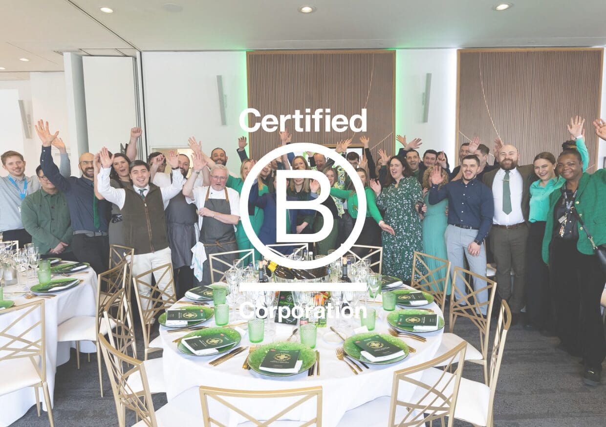 A diverse group of people celebrate around a table set for a formal event. Many have their arms raised in excitement. A large “Certified B Corporation” logo is superimposed over the image.