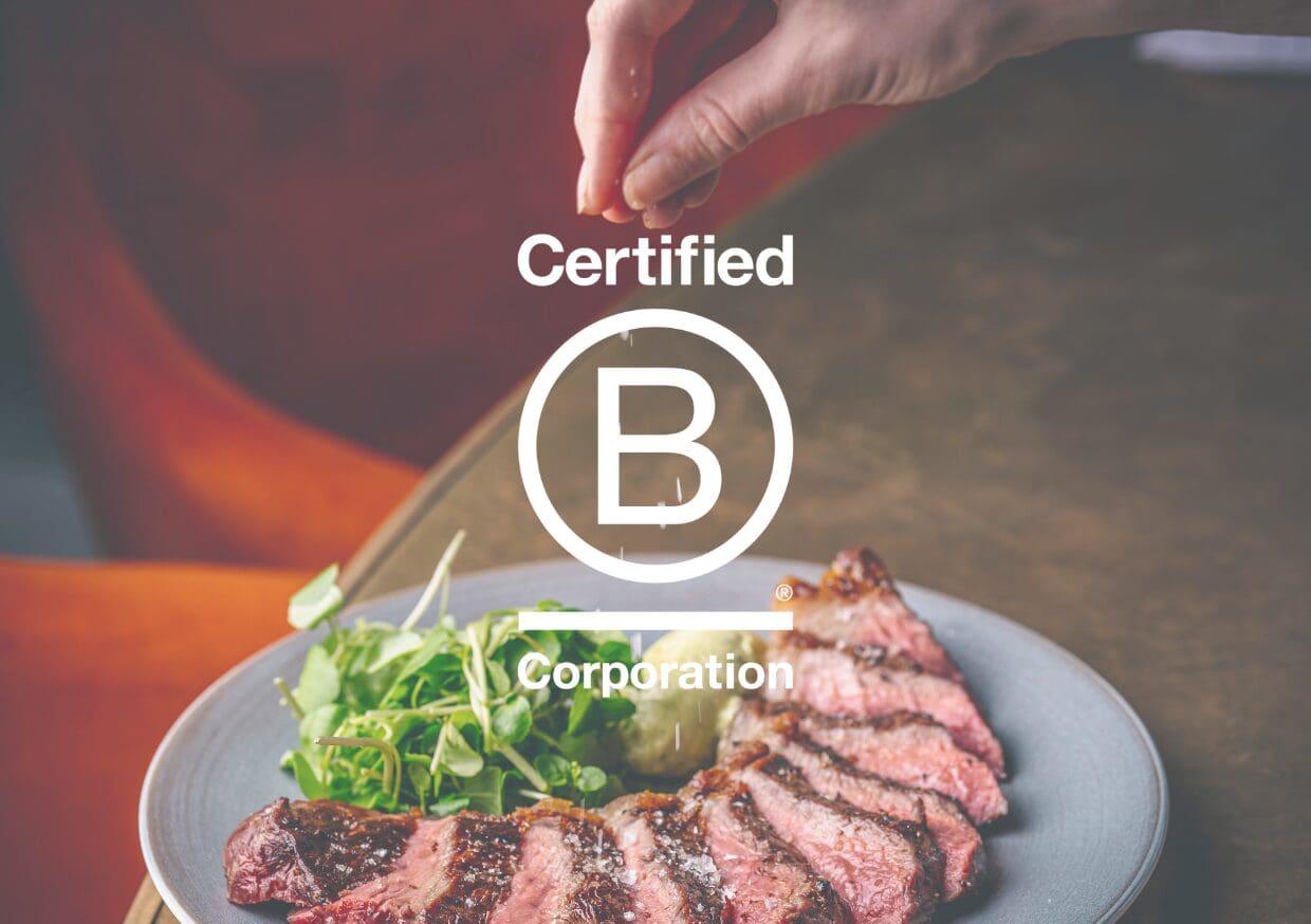 A hand sprinkles salt over sliced steak and greens on a plate, with the Certified B Corporation logo overlaid in the centre of the image.
